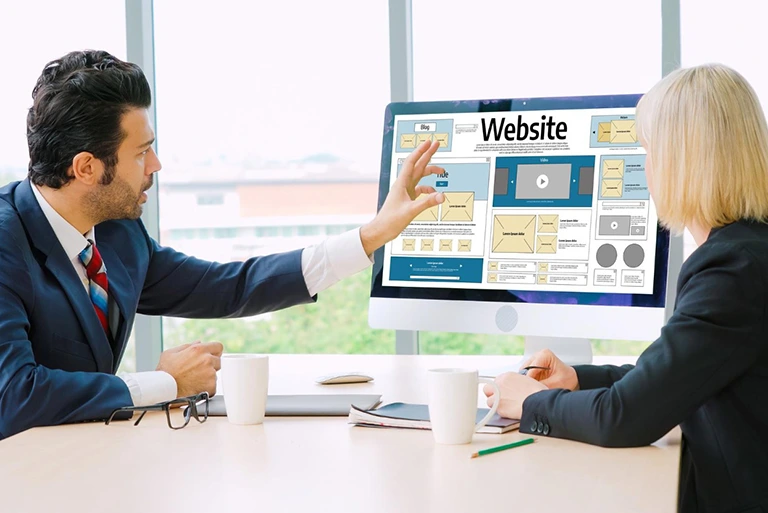 Two professionals reviewing a website layout design on a desktop computer during a business meeting.